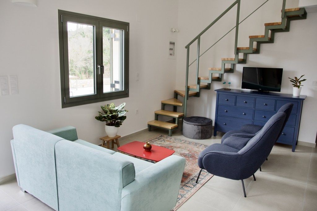 Modern living room with a blue couch and a gray armchair, featuring a red coffee table and a decorative rug. A staircase with wooden steps leads to the upper level. There is a television on a blue dresser, a potted plant on a side table, and a window with a view of the outdoors.