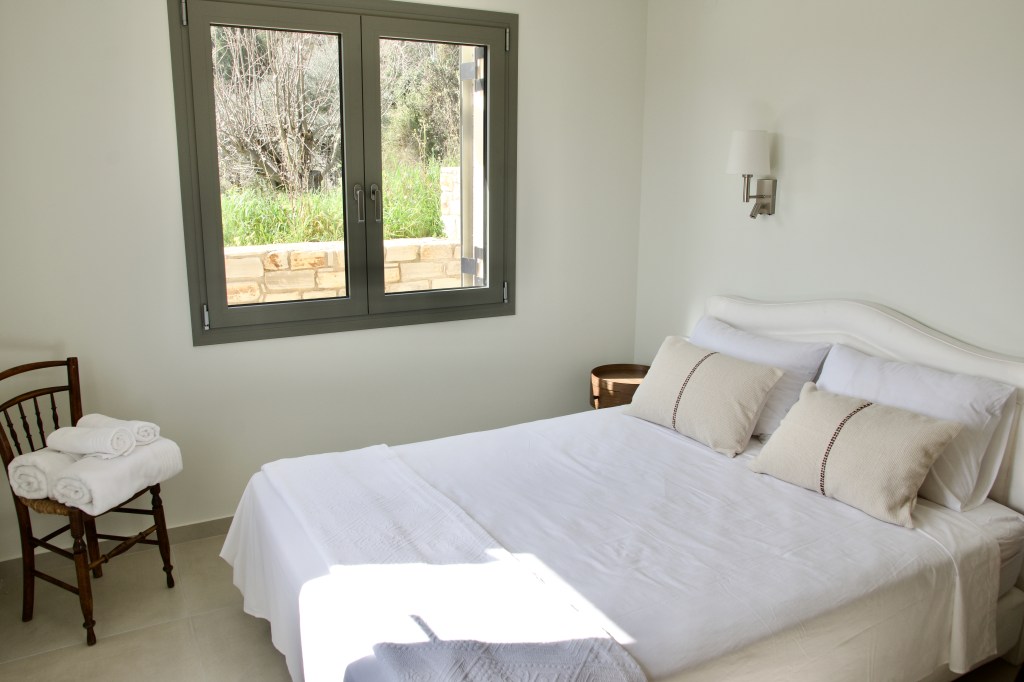 A bright and airy bedroom featuring a double bed with white linens and decorative pillows, a wooden chair with rolled towels, and a small round table. Large windows provide natural light and a view of greenery outside.